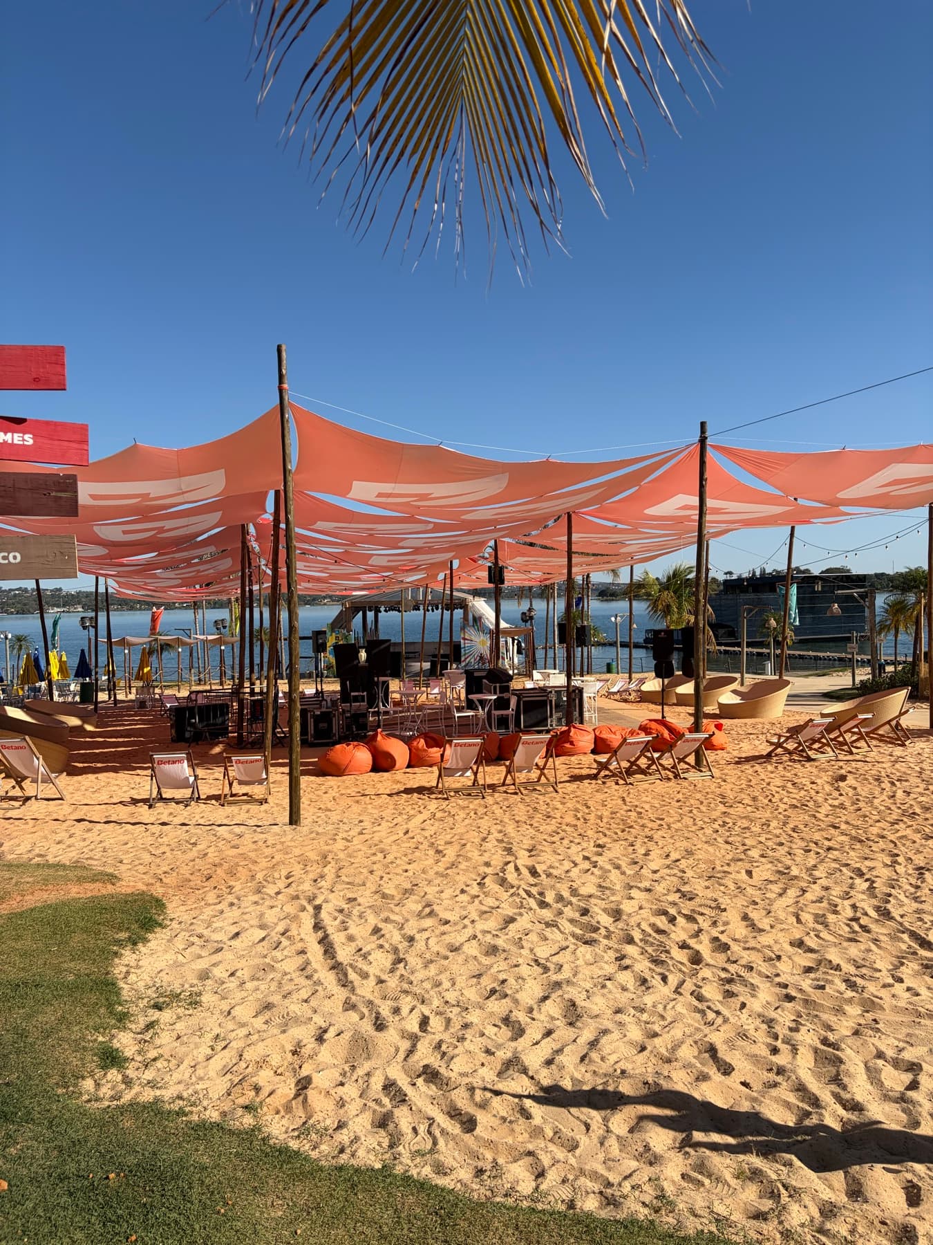 Areia, tendas coral e cadeiras de praia do Na Praia com o Lago Paranoá ao fundo